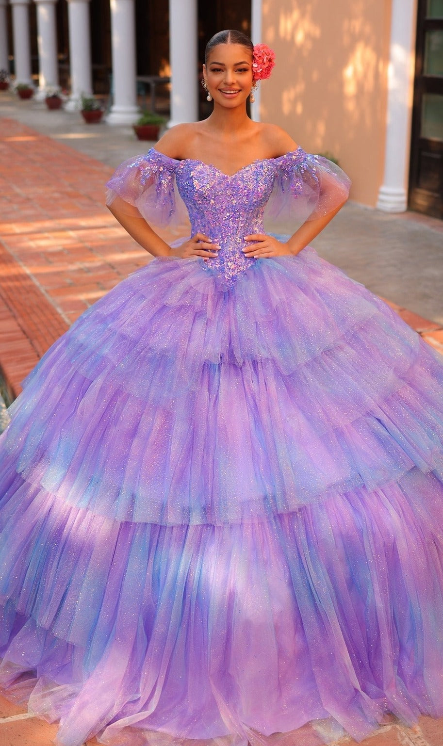 A young woman stands outdoors wearing the Quinceanera Dress 54312 by Amarra, a sparkly off-the-shoulder ball gown featuring layered tulle, a sweetheart neckline, and a pink flower in her hair.